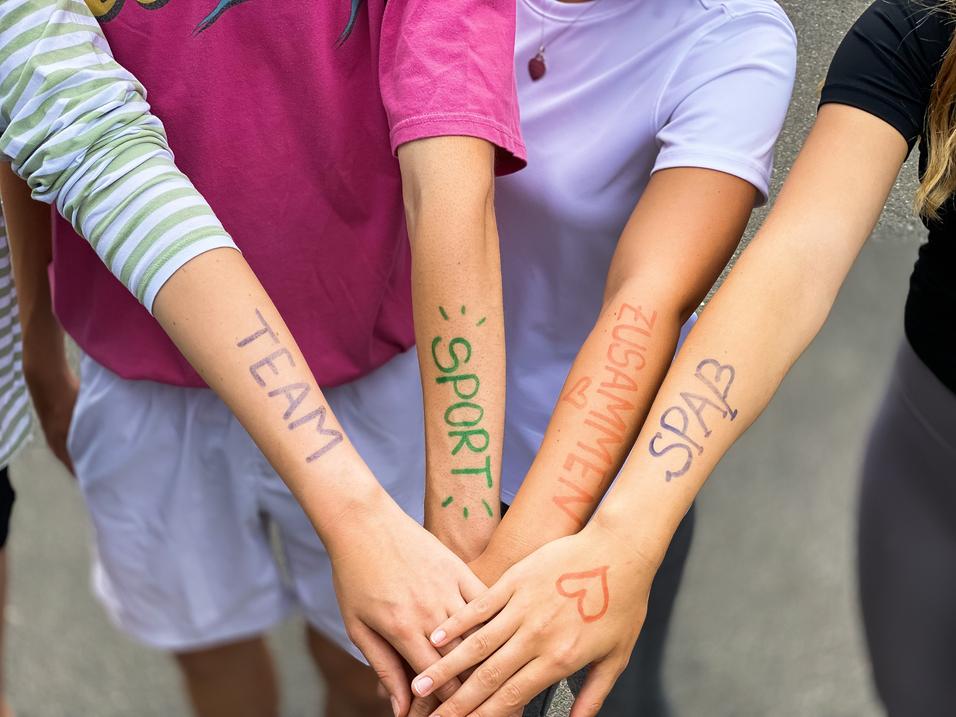 Vier Arme liegen übereinander, mit farbiger Schrift: „TEAM“, „SPORT“, „ZUSAMMEN“, „SPASS“ auf der Haut.