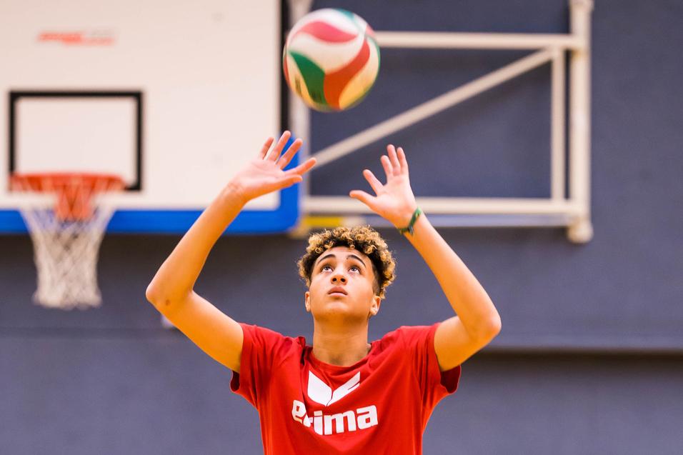 Junger Volleyballspieler hebt den Ball zum Spielen über den Kopf, in einer Sporthalle mit Basketballkorb im Hintergrund.